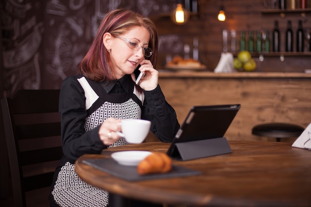 Imprenditrice adulta di successo che parla al telefono mentre tiene in mano una tazza di caffè. Ristorante vintage