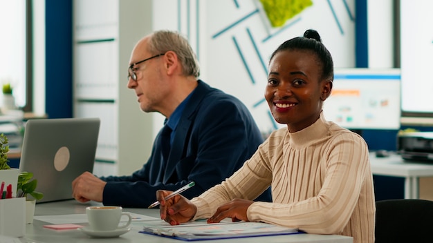 Imprenditrice africana analizzando il rapporto e guardando la telecamera sorridente seduta alla scrivania della conferenza durante il brainstorming. Imprenditore che lavora in un'attività finanziaria di avvio professionale pronto per l'incontro