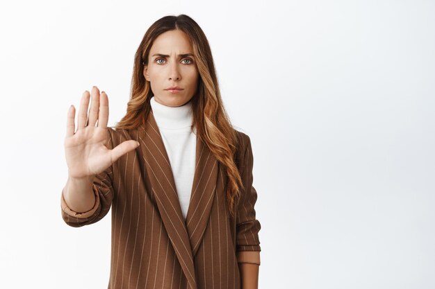 Imprenditrice arrabbiata che allunga il palmo della mano mostrando stop nessun gesto di proibizione accigliata delusa in piedi su sfondo bianco