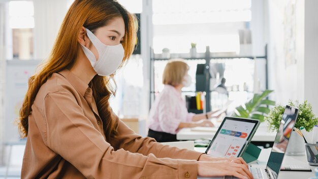 Imprenditrice asiatica che indossa una maschera medica per il distanziamento sociale in una nuova situazione normale per la prevenzione dei virus mentre si utilizza il laptop al lavoro in ufficio.