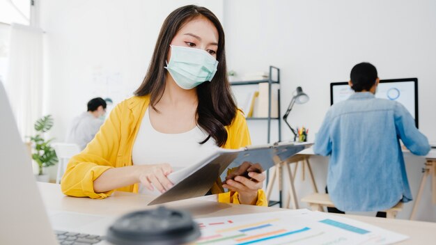 Imprenditrice asiatica imprenditrice indossando maschera medica per l'allontanamento sociale in una nuova situazione normale per la prevenzione dei virus mentre si utilizza il laptop al lavoro in ufficio. Stile di vita dopo il virus corona.