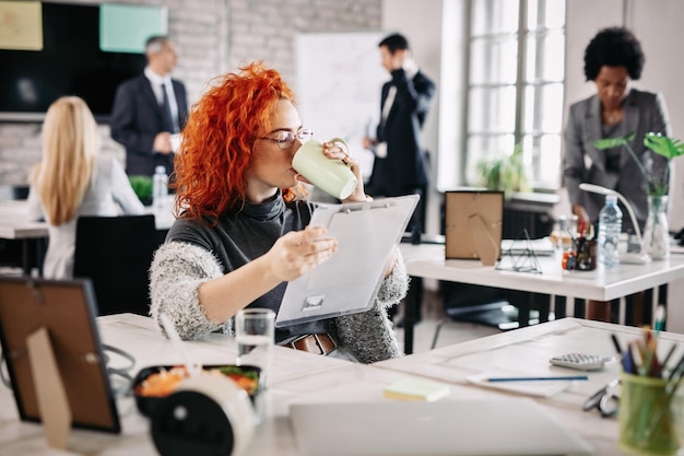 Imprenditrice che legge rapporti aziendali e beve caffè mentre è seduta alla sua scrivania in ufficio Ci sono persone sullo sfondo