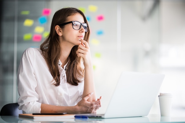 Imprenditrice di successo che lavora al computer portatile e pensa a nuove idee nel suo ufficio vestito con abiti bianchi