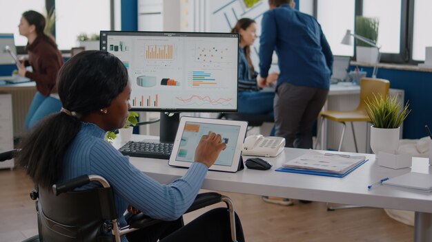 Imprenditrice disabile con disabilità locomotorie che utilizza computer e tablet allo stesso tempo lavorando in un ufficio di avviamento aziendale