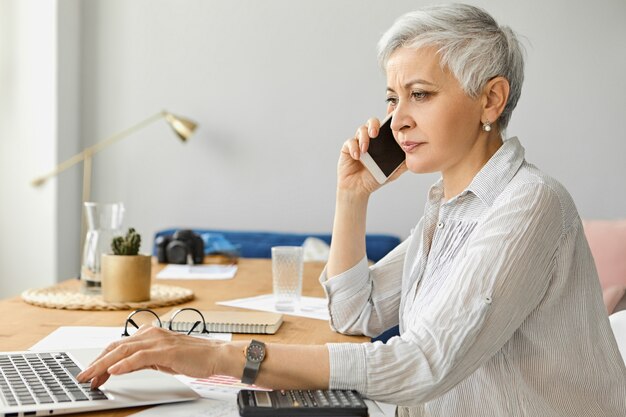 Imprenditrice matura fiduciosa di successo con i capelli corti grigi che lavora all'interno dell'ufficio elegante, utilizzando laptop e calcolatrice, parlando con il partner commerciale tramite telefono cellulare. Persone, età e occupazione