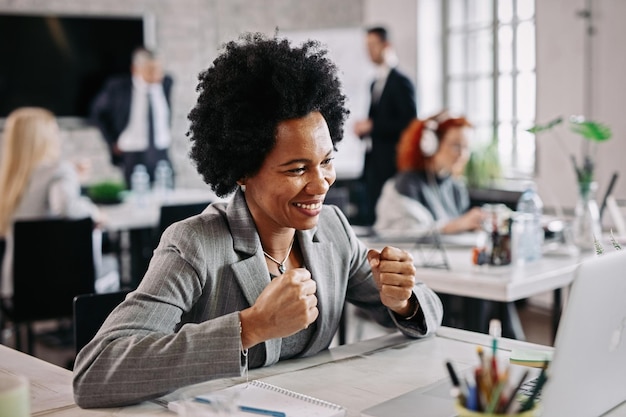 Imprenditrice nera di successo che lavora su un computer e festeggia in ufficio Ci sono persone in background