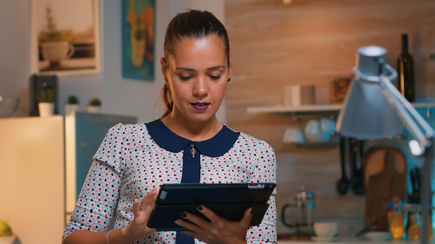 Imprenditrice prendendo pausa utilizzando tablet a tarda notte seduto in cucina moderna. Impiegato impegnato e concentrato che utilizza la moderna tecnologia di rete wireless facendo gli straordinari, scrivendo, cercando.