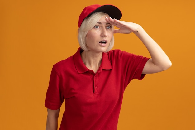 Impressionata donna bionda di mezza età in uniforme rossa e berretto che tiene la mano sulla fronte guardando di lato a distanza