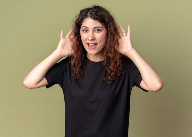 Impressionata giovane bella donna che guarda davanti facendo grandi orecchie isolate su un muro verde oliva