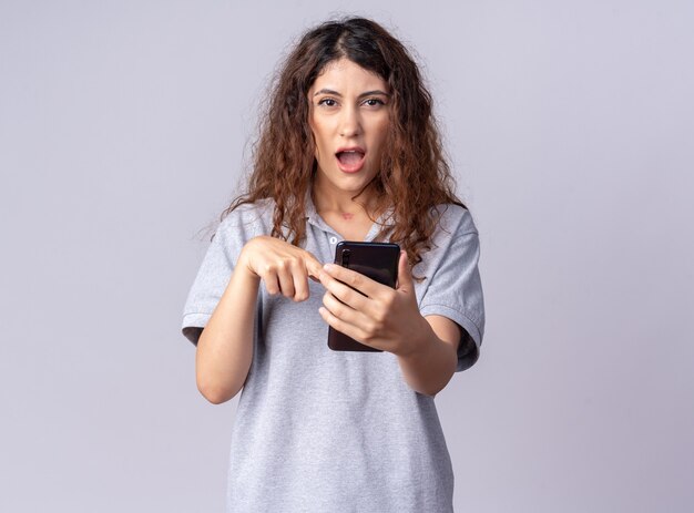 Impressionata giovane bella donna che tiene e indica il telefono cellulare guardando la parte anteriore isolata sul muro bianco con copia spazio