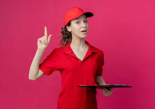 Impressionata giovane bella ragazza delle consegne in uniforme rossa e berretto che tiene appunti e alzando il dito sullo spazio cremisi