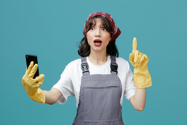 Impressionata giovane donna addetta alle pulizie che indossa bandana uniforme e guanti di gomma che allungano il telefono cellulare guardando il lato rivolto verso l'alto isolato su sfondo blu