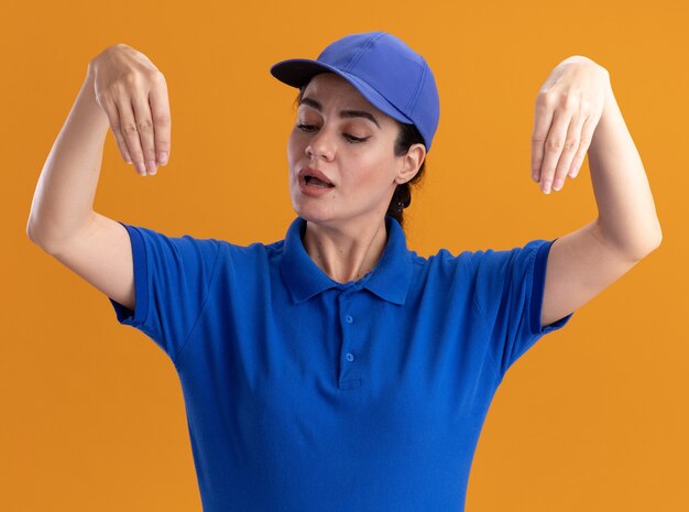 Impressionata giovane donna delle consegne in uniforme e berretto finge di tenere qualcosa di fronte a lei guardandolo isolato sul muro arancione