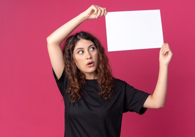 Impressionata giovane donna graziosa che tiene carta bianca sopra la testa guardandola isolata sul muro cremisi