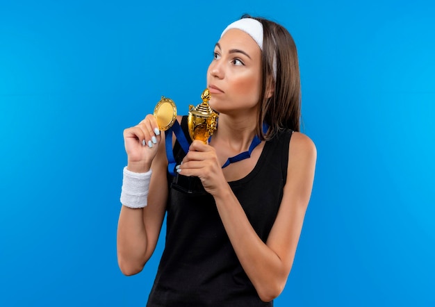 Impressionata giovane ragazza abbastanza sportiva che indossa fascia e cinturino e medaglia al collo tenendo la coppa e la medaglia del vincitore e guardando il lato isolato sulla parete blu