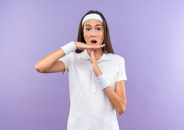 Impressionata ragazza piuttosto sportiva che indossa fascia e cinturino che gesturing timeout isolato sul muro viola