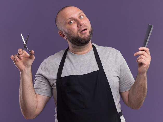 Impressionato barbiere maschio di mezza età in uniforme che tiene le forbici con pettine isolato su parete viola