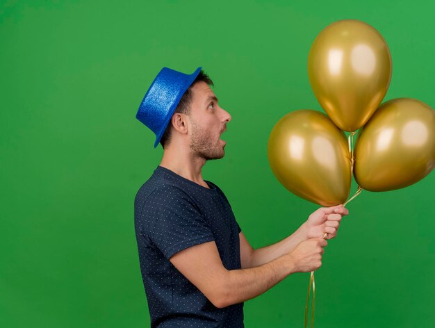 Impressionato bell'uomo che indossa il cappello blu del partito si leva in piedi lateralmente guardando e tenendo palloncini di elio isolati sulla parete verde con lo spazio della copia