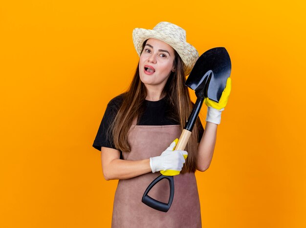 Impressionato bella ragazza giardiniere che indossa uniforme e cappello da giardinaggio con guanti che tengono vanga isolato su sfondo arancione