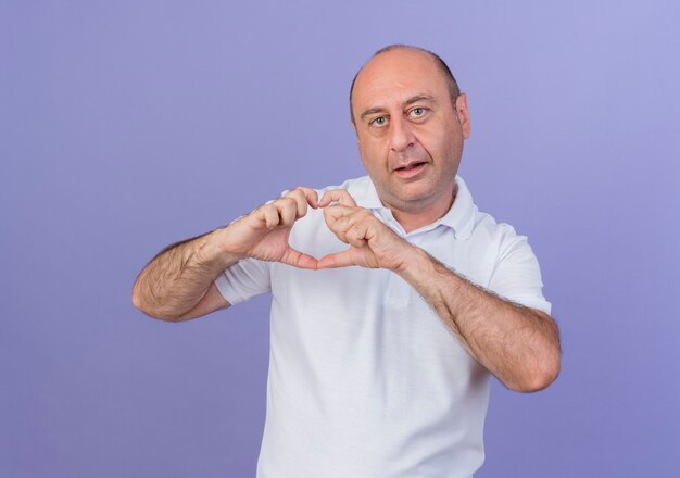 Impressionato casual uomo d'affari maturo guardando la fotocamera e facendo segno di cuore isolato su sfondo viola