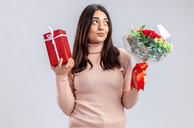 Impressionato cercando giovane ragazza il giorno di San Valentino con scatola regalo con bouquet isolato su sfondo bianco