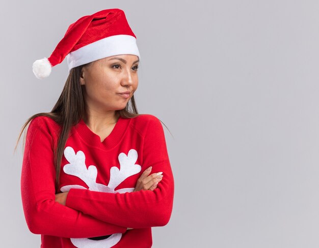 Impressionato cercando lato giovane ragazza asiatica che indossa il cappello di Natale con le mani incrociate maglione isolato su sfondo bianco con spazio di copia