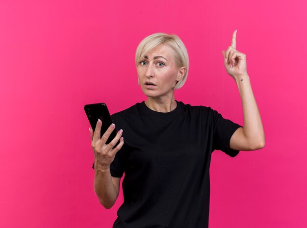 Impressionato di mezza età bionda donna slava tenendo il telefono cellulare guardando la telecamera rivolta verso l'alto isolato su sfondo cremisi con lo spazio della copia