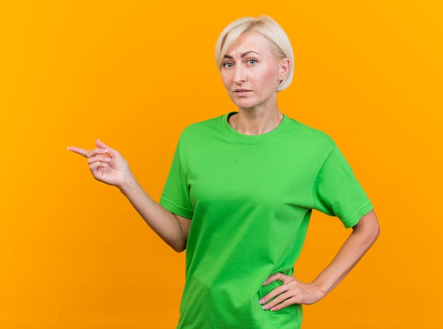 Impressionato donna bionda di mezza età guardando davanti tenendo la mano sulla vita che punta sul lato isolato sulla parete gialla