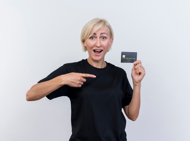 Impressionato donna slava bionda di mezza età che mostra la carta di credito che punta a esso guardando la telecamera isolata su sfondo bianco