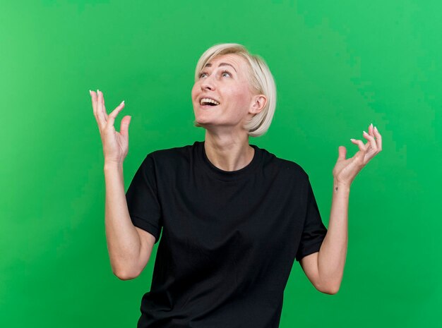Impressionato donna slava bionda di mezza età tenendo le mani in aria cercando isolato sulla parete verde