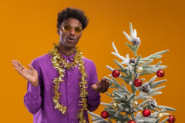 Impressionato giovane afro-americano con gli occhiali con la ghirlanda di orpelli intorno al collo in piedi vicino all'albero di Natale decorato su sfondo arancione