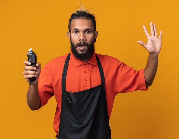 Impressionato giovane barbiere afroamericano che indossa l'uniforme che tiene i tagliacapelli che mostrano cinque con la mano