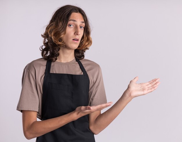 Impressionato giovane barbiere bello che indossa l'uniforme che punta a lato con le mani isolate sul muro bianco