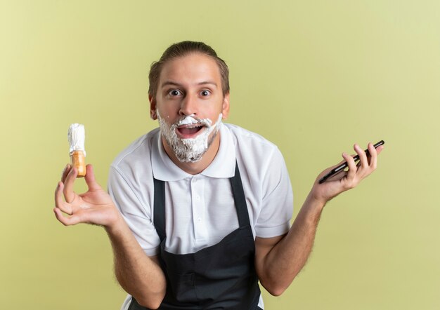 Impressionato giovane barbiere bello che indossa l'uniforme che tiene il telefono cellulare e il pennello da barba con crema da barba applicata sulla sua barba isolata su verde oliva