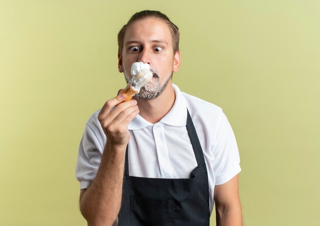 Impressionato giovane barbiere bello che indossa l'uniforme guardando il pennello da barba con crema da barba applicata sul suo viso isolato su verde oliva con spazio di copia