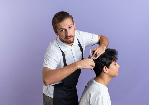 Impressionato giovane barbiere bello che indossa uniforme facendo taglio di capelli per il giovane cliente isolato su viola con spazio di copia