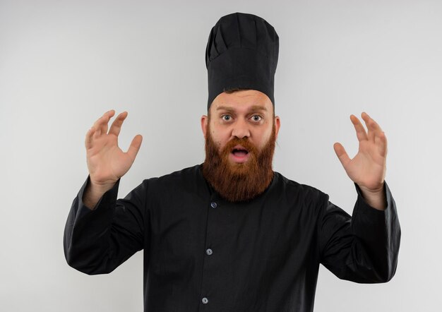 Impressionato giovane bel cuoco in uniforme da chef alzando le mani isolato su muro bianco