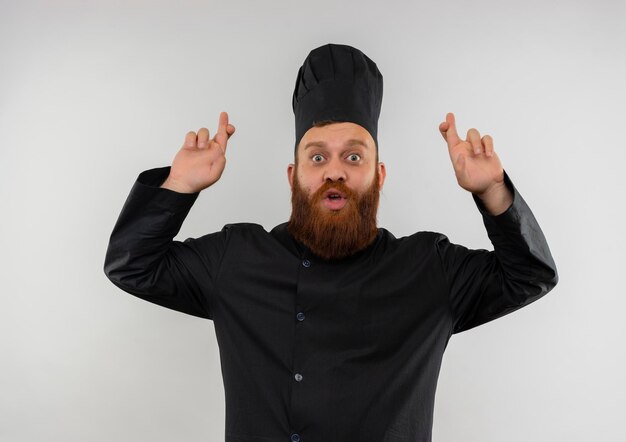 Impressionato giovane bel cuoco in uniforme da chef che fa gesto di dita incrociate isolato su muro bianco