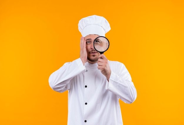 Impressionato giovane bel cuoco in uniforme da chef guardando attraverso la lente d'ingrandimento con la mano sul viso sulla parete arancione isolata con spazio copia copy