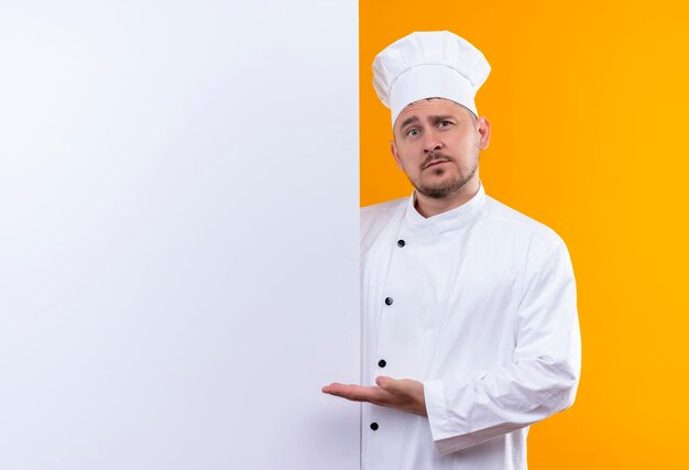 Impressionato giovane bel cuoco in uniforme da chef in piedi dietro il muro bianco e indicandolo isolato sul muro arancione con spazio copia