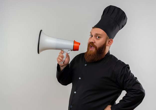 Impressionato giovane bel cuoco in uniforme da chef parlando da un altoparlante isolato su muro bianco white