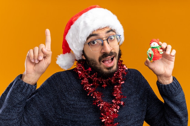 Impressionato giovane bel ragazzo che indossa il cappello di Natale con la ghirlanda sul collo che tiene i punti del giocattolo in alto isolato sulla parete arancione