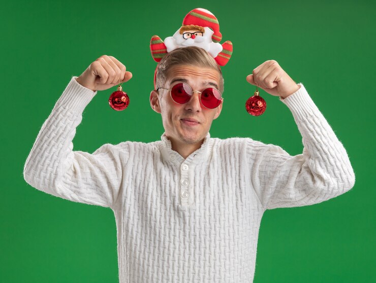 Foto gratuito impressionato giovane bel ragazzo che indossa la fascia di babbo natale con gli occhiali che tengono le bagattelle di natale che guarda l'obbiettivo isolato su priorità bassa verde
