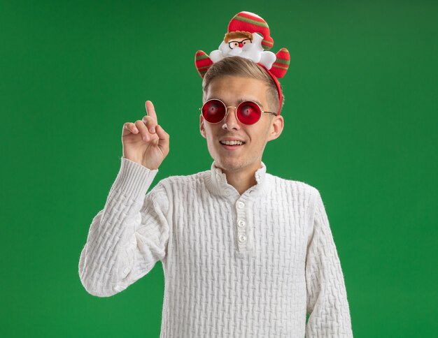 Impressionato giovane bel ragazzo che indossa la fascia di Babbo Natale con gli occhiali guardando a lato e rivolto verso l'alto isolato su sfondo verde