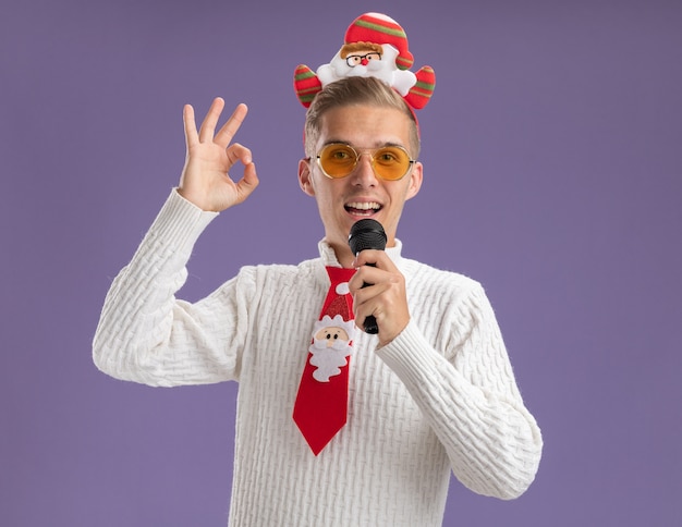 Impressionato giovane bel ragazzo che indossa la fascia di Babbo Natale e cravatta con gli occhiali che tiene il microfono vicino alla bocca facendo segno ok isolato sulla parete viola