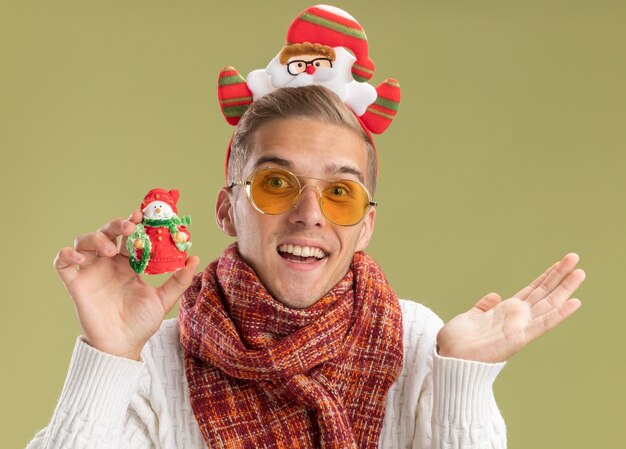 Impressionato giovane bel ragazzo che indossa la fascia di Babbo Natale e sciarpa che tiene l'ornamento di Natale del pupazzo di neve che mostra la mano vuota isolata sulla parete verde oliva