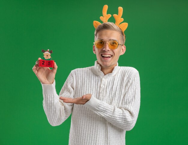 Impressionato giovane bel ragazzo che indossa la fascia di corna di renna con gli occhiali che tengono il giocattolo dell'albero di Natale con la data che indica con la mano isolata sulla parete verde con lo spazio della copia