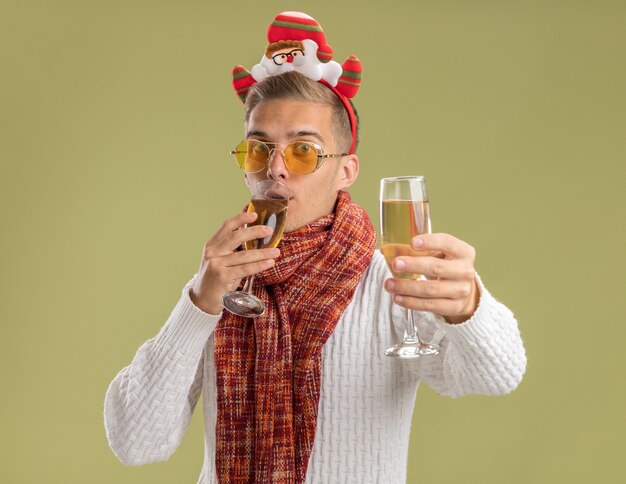 impressionato giovane bel ragazzo che indossa la fascia e la sciarpa di babbo natale guardando tenendo in mano due bicchieri di champagne bevendone uno e allungandone un altro verso la telecamera isolata sul muro verde oliva