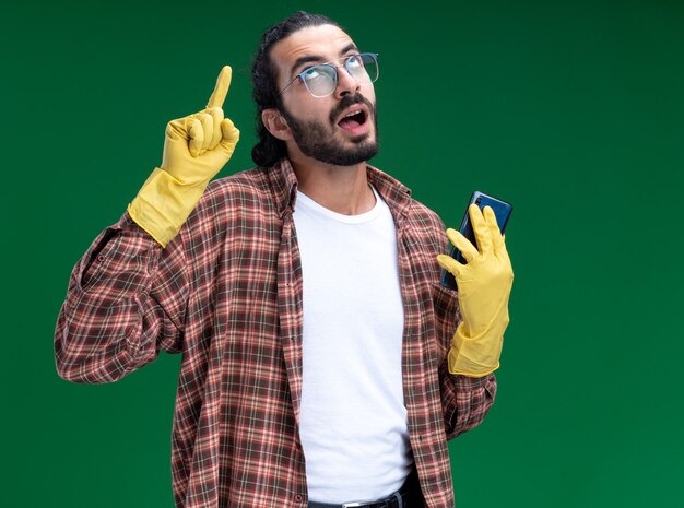 Impressionato giovane bel ragazzo delle pulizie che indossa t-shirt e guanti che tengono i punti del telefono in alto isolati sul muro verde