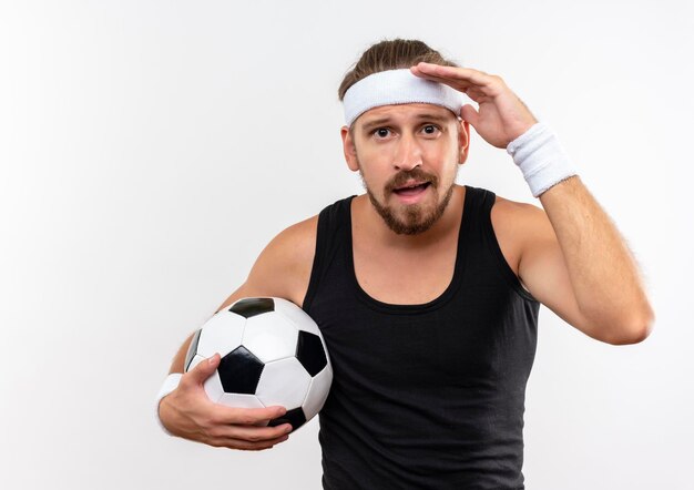 Impressionato giovane bell'uomo sportivo che indossa fascia e braccialetti che tengono in mano un pallone da calcio e che mettono la mano vicino alla fronte isolata sul muro bianco